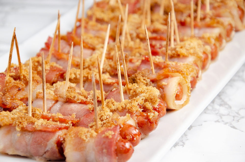 Bacon wrapped smokies with brown sugar lined up on a white plate with marble table underneath.