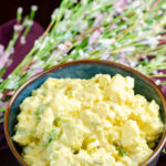 Deviled egg potato salad in blue bowl. Spring flowers and purple napkin in background.