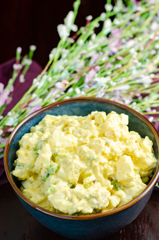 Deviled egg potato salad in blue bowl. Spring flowers and purple napkin in background.