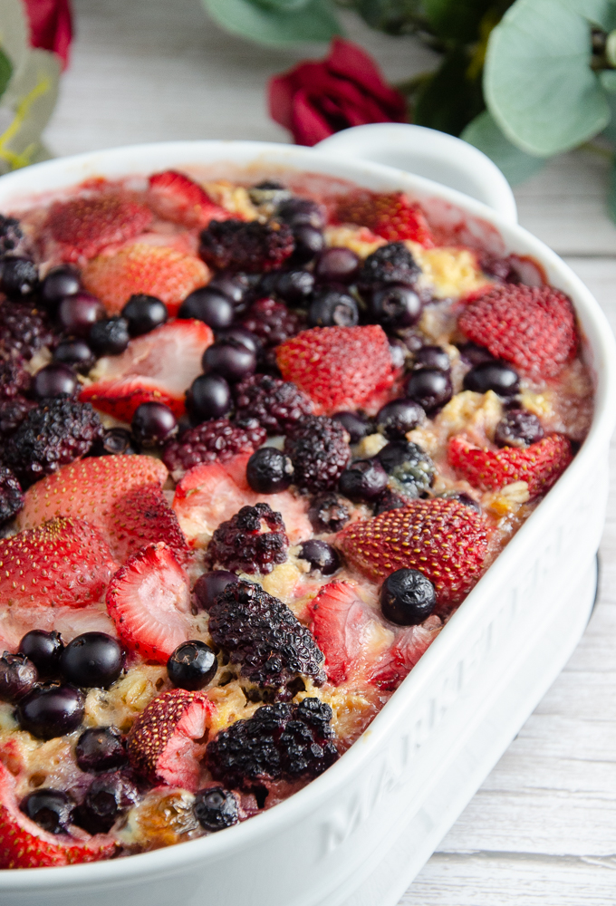 Triple berry baked oatmeal in white oval casserole dish. Rose in background. White wood for floor.
