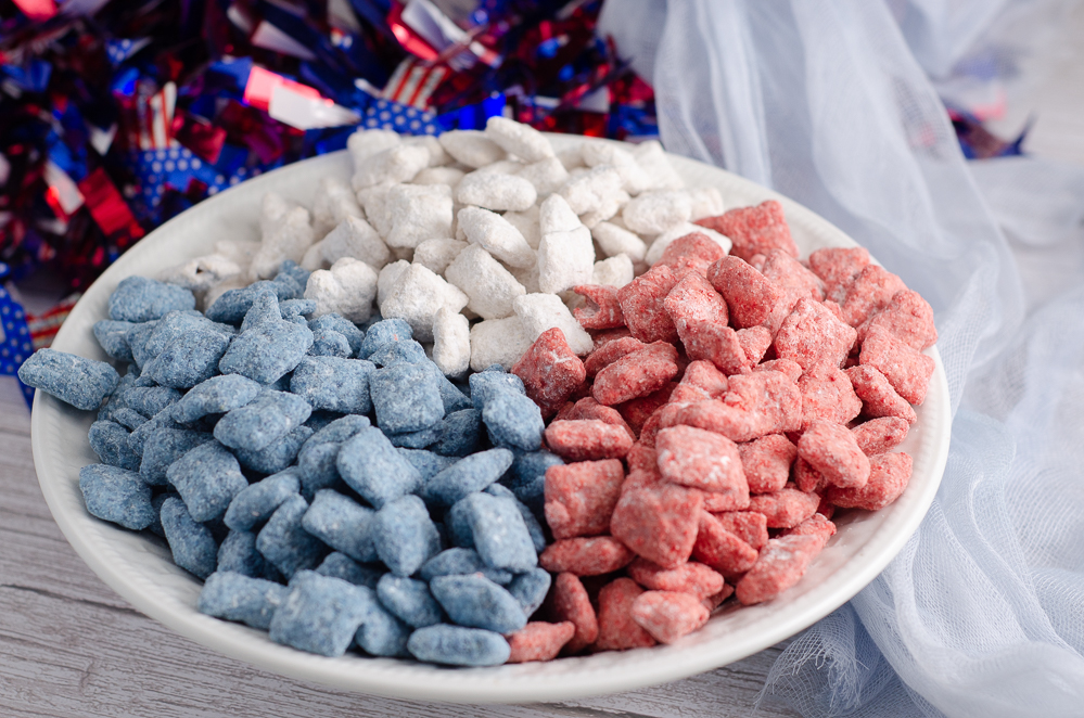 Patriotic muddy buddies- divided by color- in white round dish with sparkle wreath in background.