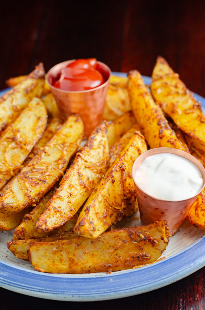 Kickin' baked parmesan potato wedges, vertical picture. Piles on plate with cups of sour cream and ketchup.