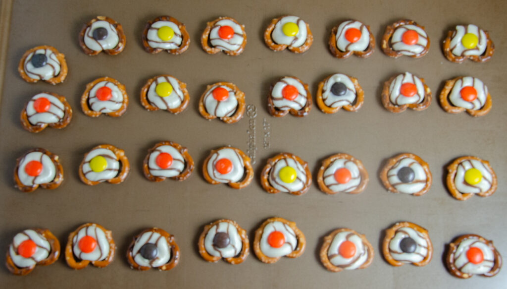 Cookie sheet with pretzels. The striped Hershey's Kisses are melted and the Reese's Pieces have bene placed on top of each melted kiss.