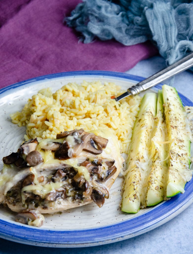 Mushroom and smoked Gouda stuffed chicken breast on white pottery plate with blue edge. Plate also contains zucchini sticks and rice pilaf.