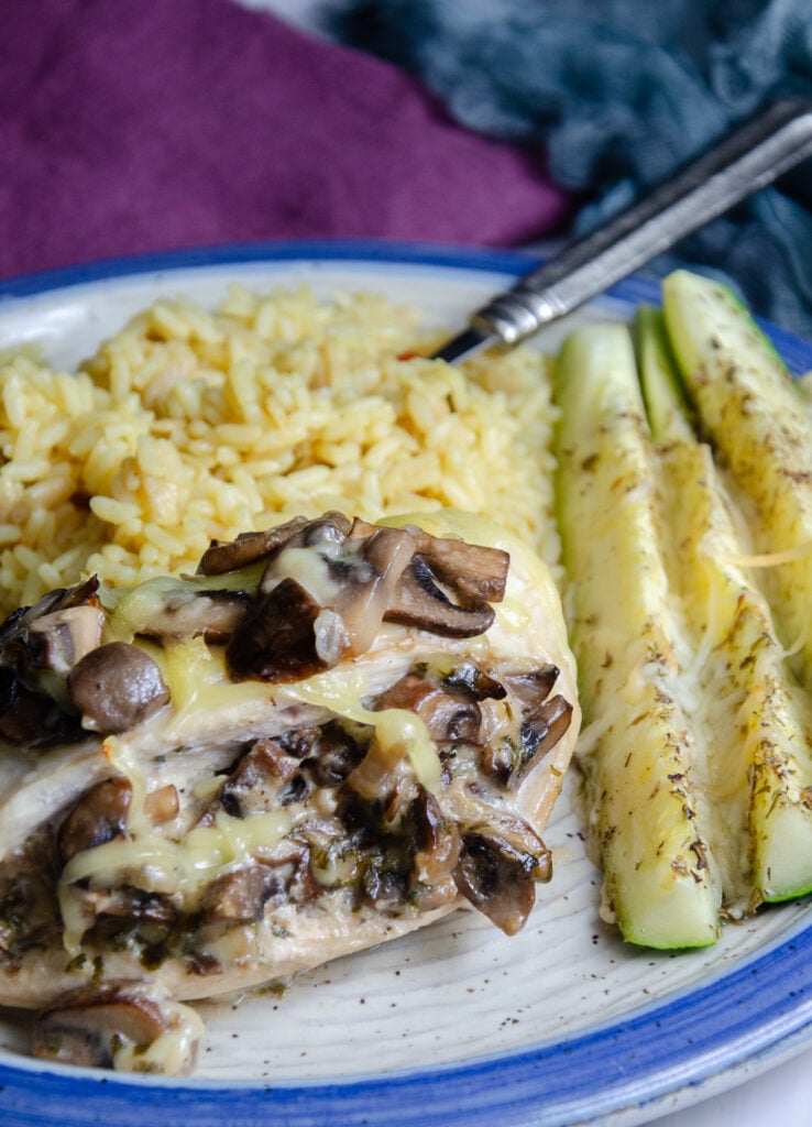 Mushroom and smoked Gouda stuffed chicken breast on white pottery plate with blue edge. Plate also contains zucchini sticks and rice pilaf.