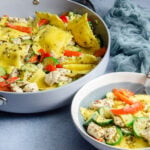 Cheese ravioli, basil pesto, chicken, zucchini, and red pepper. A large pan filled with it and a small blue and white bowl in front.