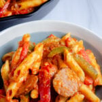 Polska kielbasa, penne pasta, bell peppers, and onion with a red sauce. In a white and grayish blue bowl with a glimpse of large black cast-iron skillet full in background.