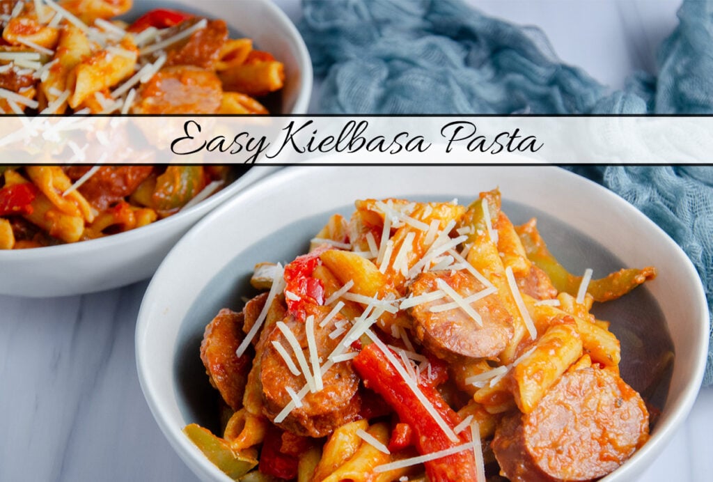 Polska kielbasa, penne pasta, bell peppers, and onion with a red sauce. Dished up in two white and grayish blue bowls on a white and gray background with light blue cheesecloth. Text overlay.